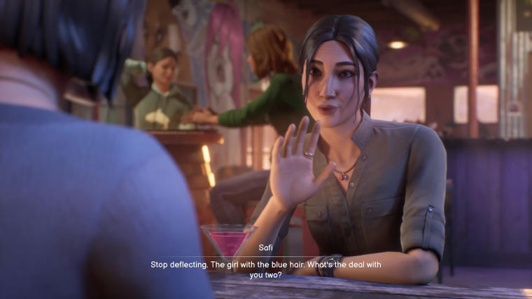 A screenshot of a scene at a bar. Max Caulfield is white, has shoulder-length dark-brow hair and a blue jumper. We see her back, not her face. Facing her at the opposite side of the table is Safi with tan skin (she's of Arabic descent) straight dark-brown hair pulled back with two strands touching her cheeks. Her elbows are on the table, her right hand raised. She's in the middle of talking to Max. The subtitles read: "Stop deflecting. The girl with the blue hair. What's the deal with you two?"